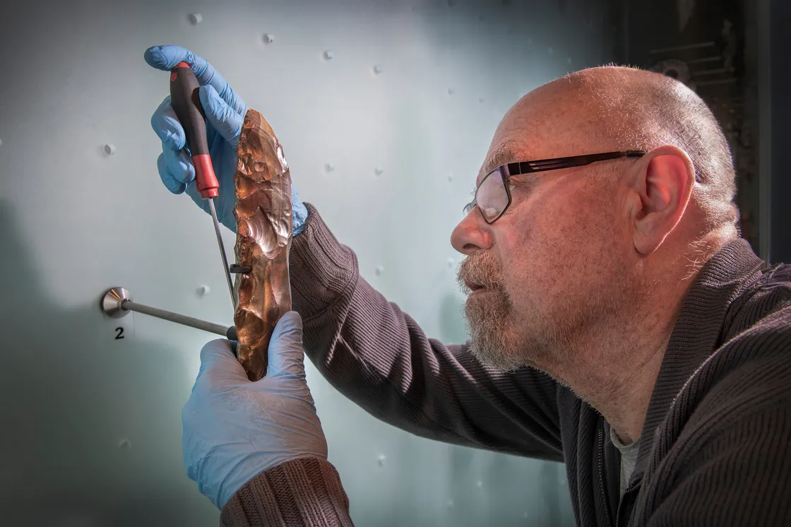 A white man wearing blue gloves removing a piece of flint from a stand
