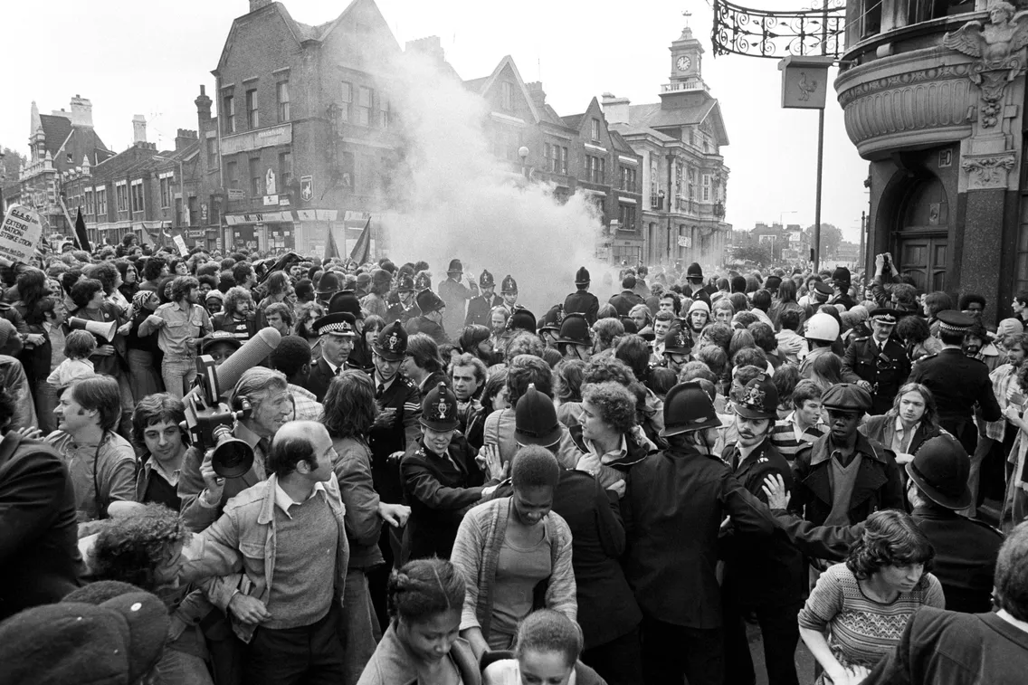 A black and white photo of a crowded street scene during a protest, with police officers and demonstrators. Smoke is visible in the background among historic buildings.