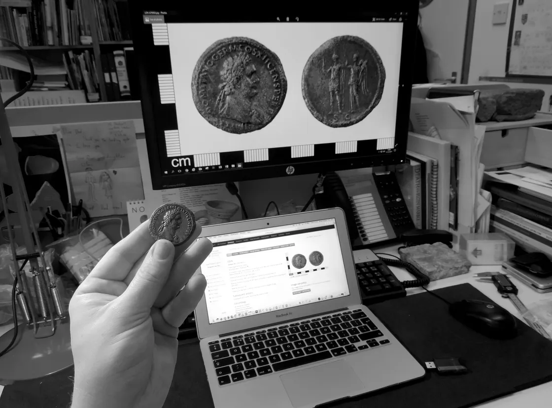 A person holding an ancient coin in their hand with its image displayed on a computer monitor, and a laptop showing research on that coin in a cluttered office.