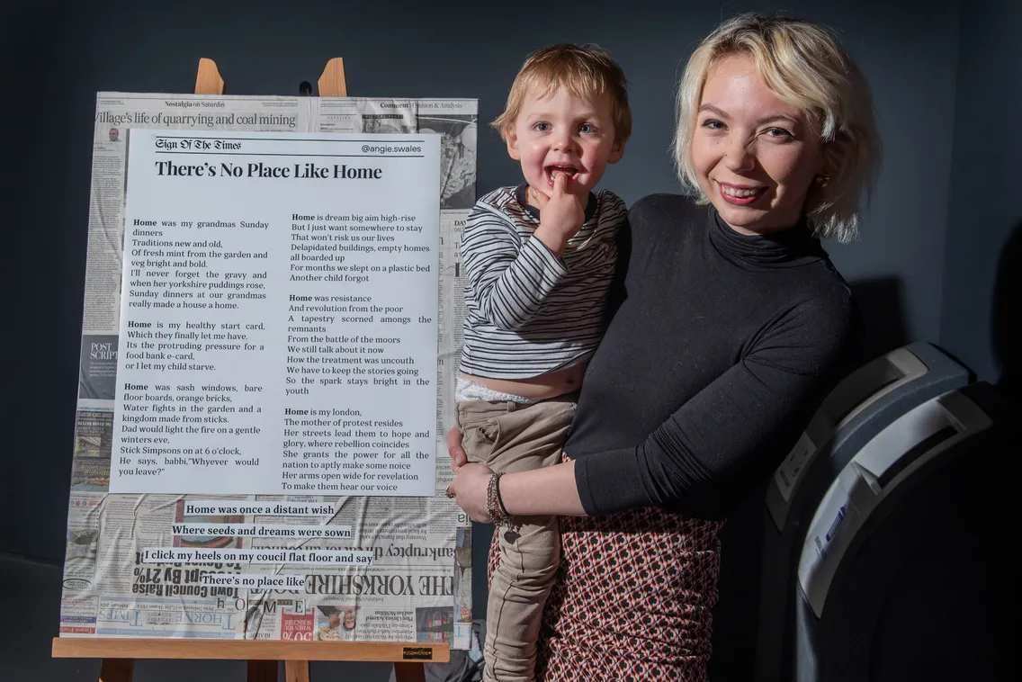 A woman holding a child smiles in front of a large poster titled "There's No Place Like Home," which is displayed on an easel and appears to be a collection of newspaper articles and clippings.