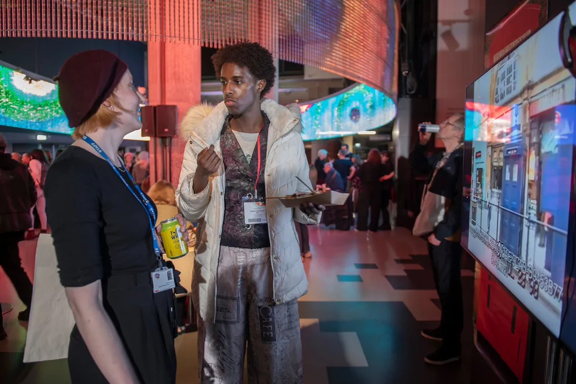 Two people are conversing at an indoor event. One is holding a drink and the other is eating from a plate. An attendee in the background is looking at a screen display.