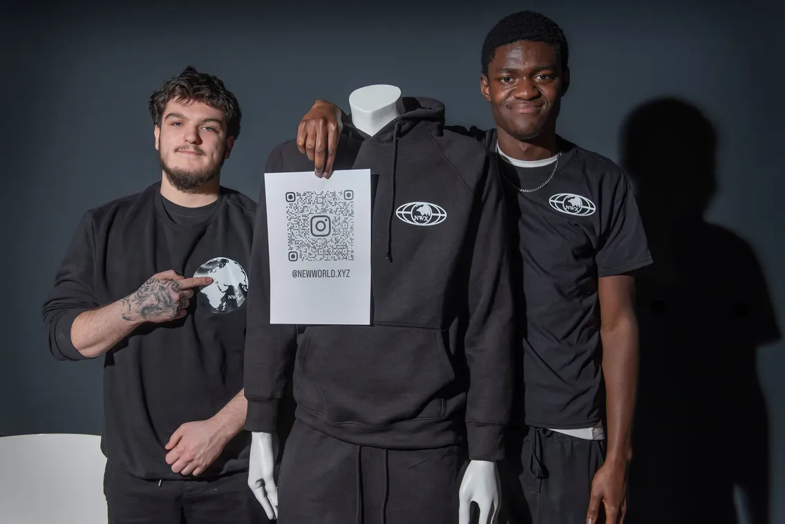Two people stand beside a mannequin wearing a black hoodie. One person points to a QR code on a paper held by the mannequin, promoting "newworld.xyz".
