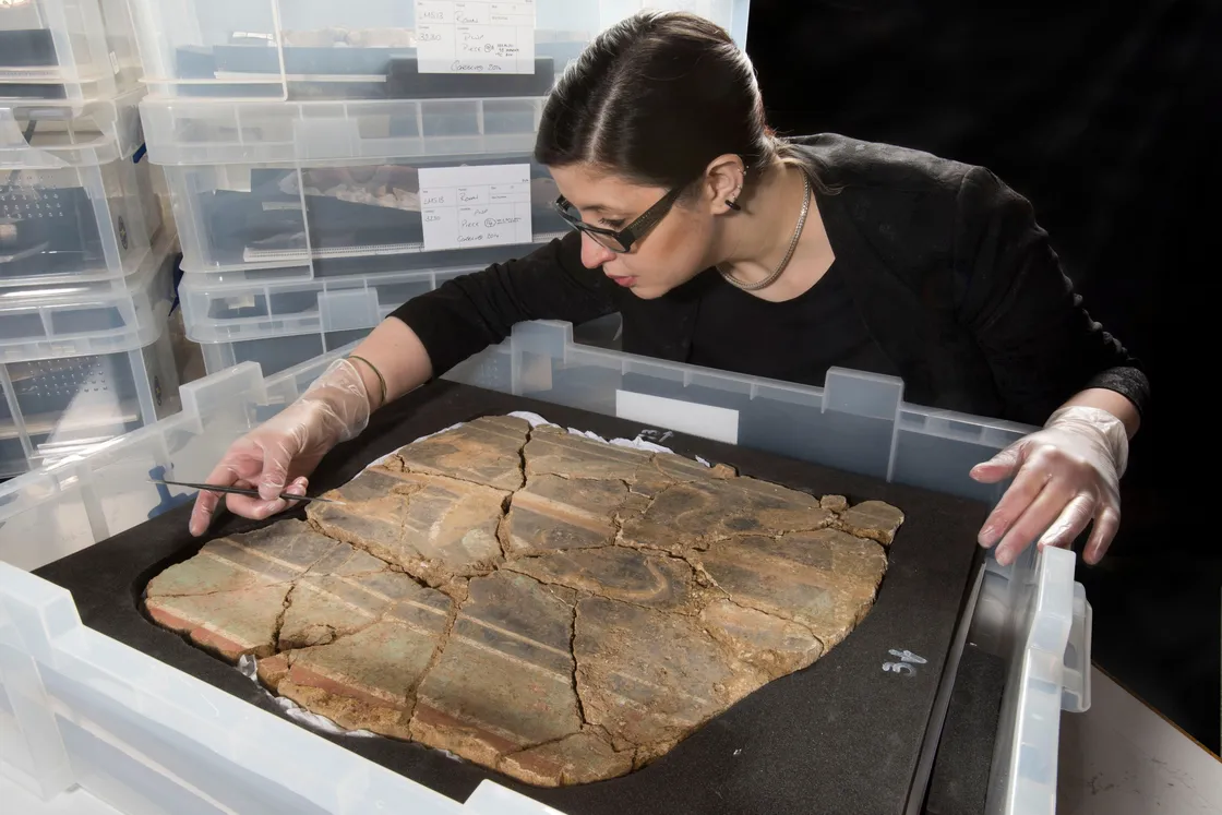 A person with glasses and green streaks in their hair is carefully examining a large, partially restored ceramic bowl at a workstation. Tools and equipment are visible in the background.