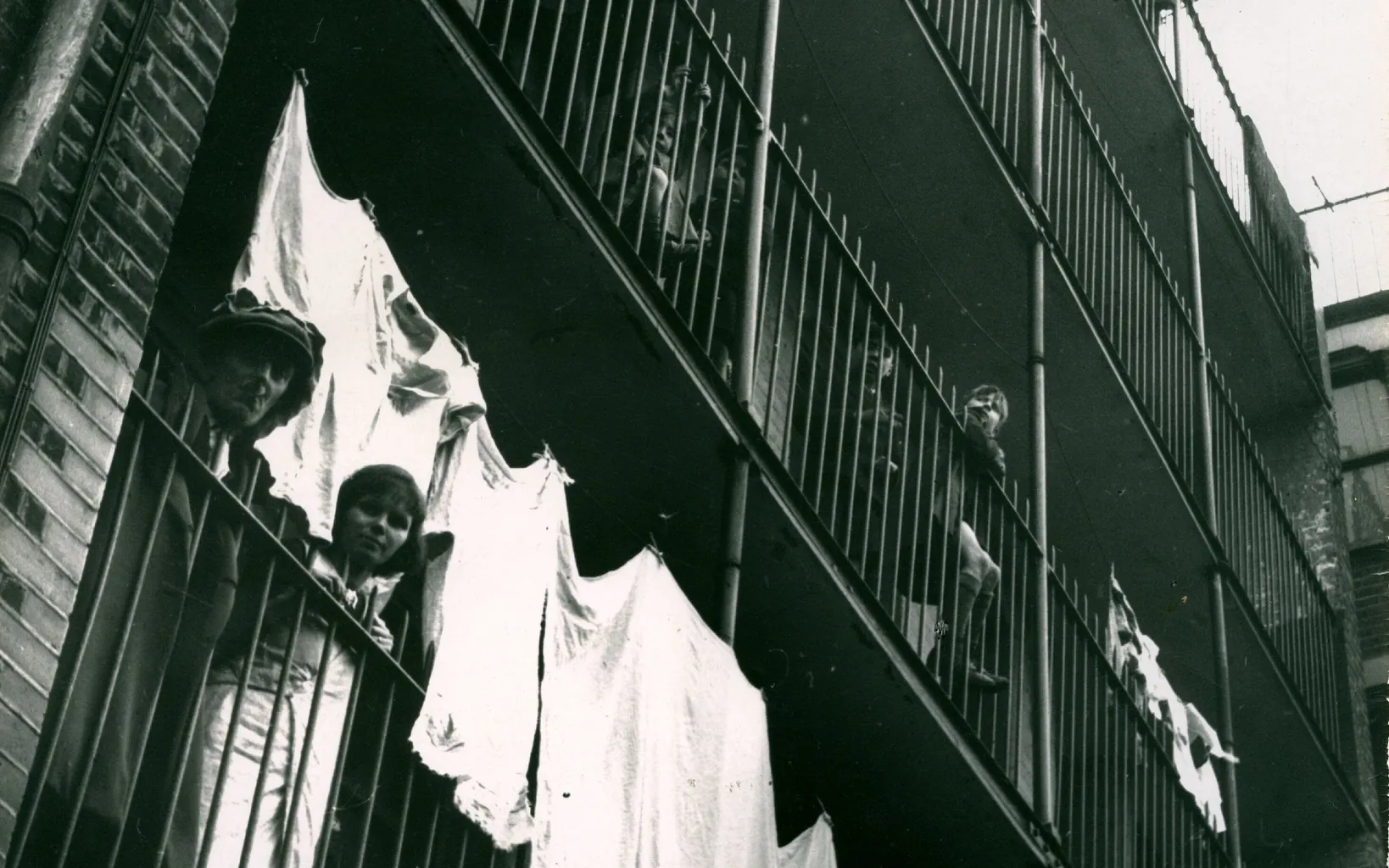 Black and white photo of residents standing on balconies of an apartment building with laundry hanging on railings.