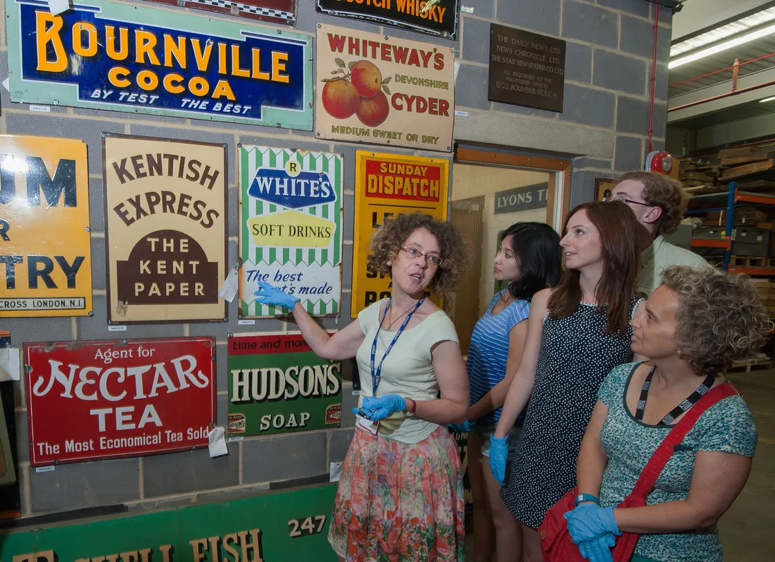 Group of five adults viewing a collection of vintage advertising signs on a wall in a museum.