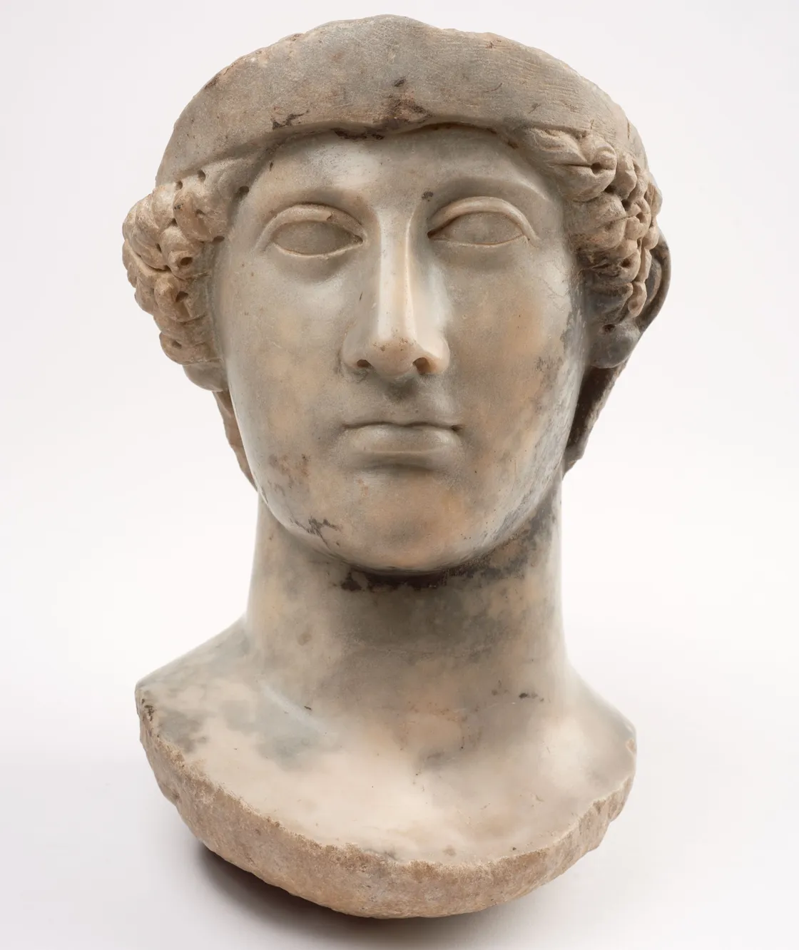A weathered marble bust of a person with a calm expression, curly hair, and a partially broken nose and neck, set against a plain white background.
