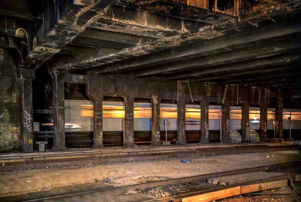 An underground railway tunnel with rusty steel beams, brick walls, and multiple tracks. A blurred train passes through in the background. Graffiti is visible on the walls.