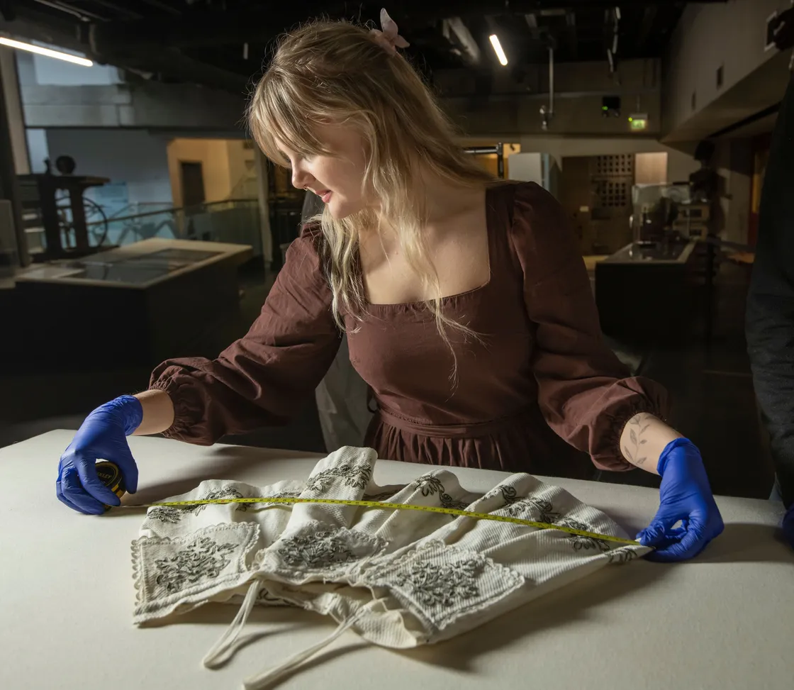 A person with long hair, wearing a brown dress and blue gloves, examines an embroidered garment on a table with a measuring tape in a dimly lit room.