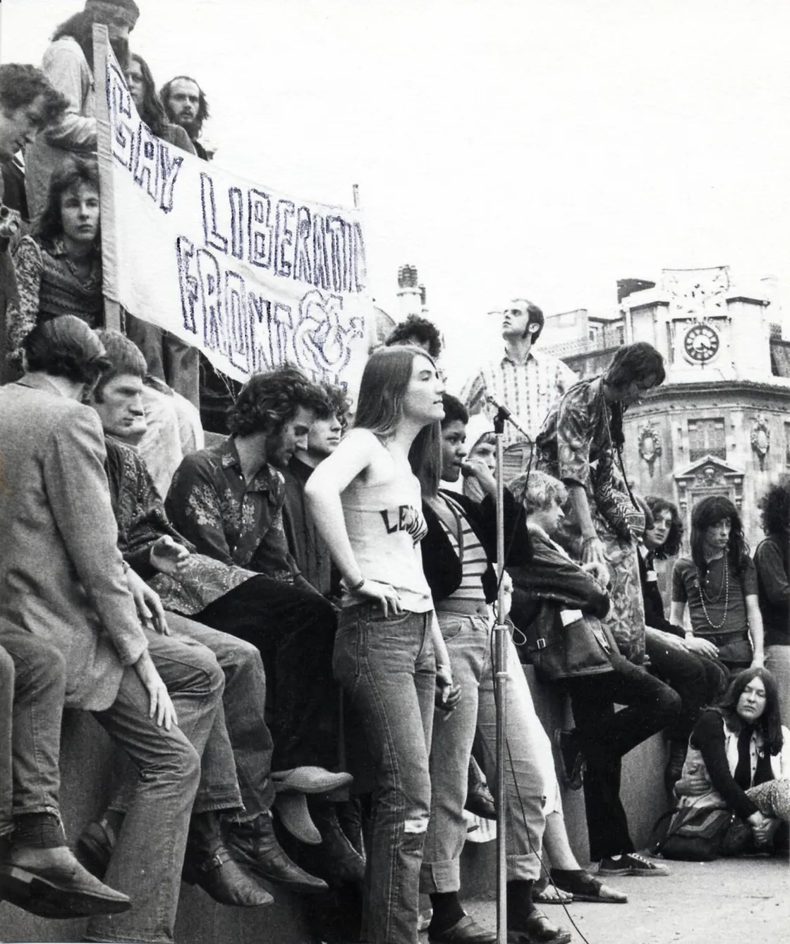 A group of people gathers outdoors near a banner reading "Gay Liberation Front" with some individuals sitting, standing, and listening to a person speaking at a microphone.