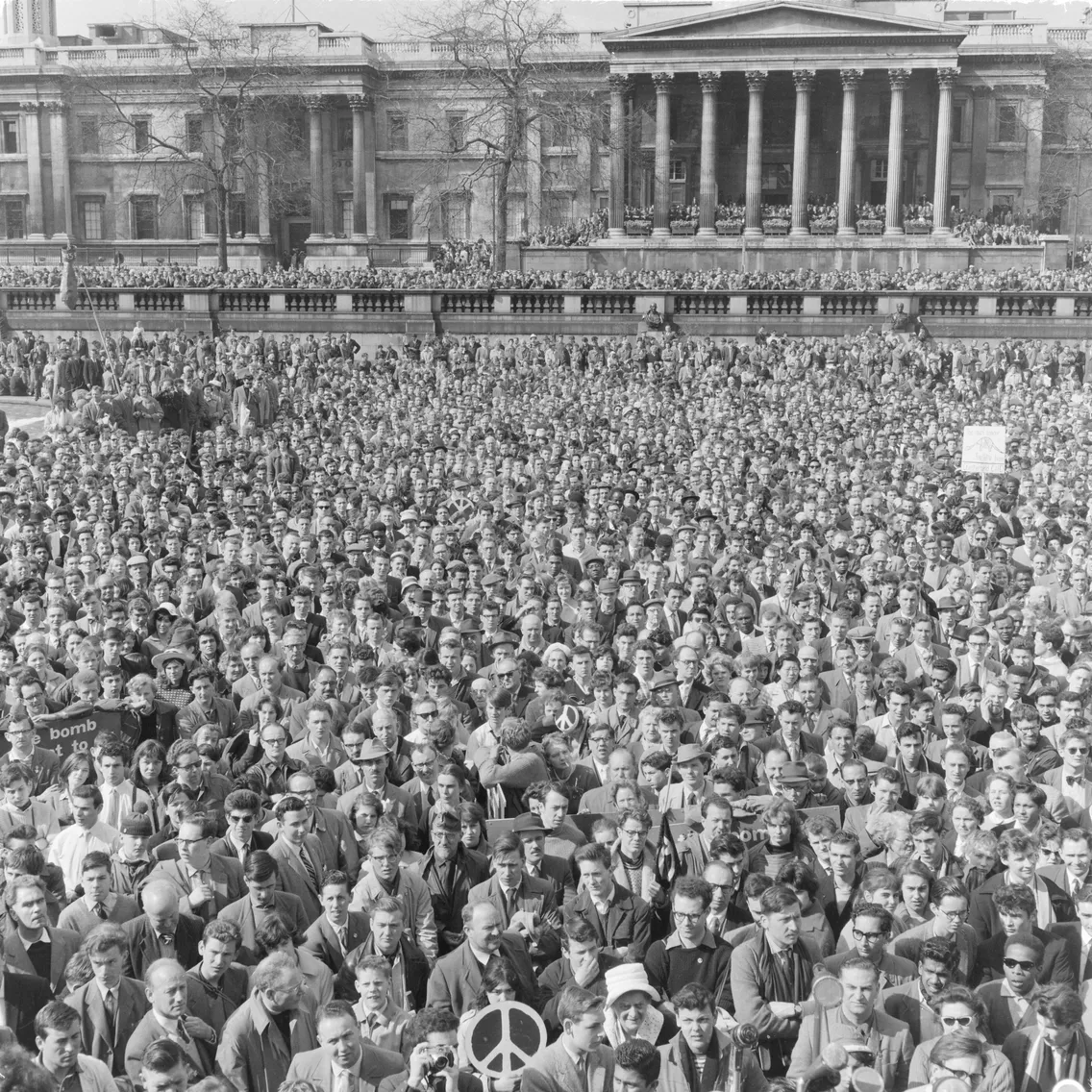 A historic black and white photo of a large crowd gathered in front of a neoclassical building, some holding peace signs during a protest.