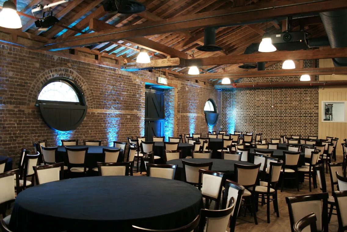 A brick-walled event space with round tables and chairs arranged, featuring arched windows and blue lighting accents. The ceiling exposes wooden beams and several light fixtures.