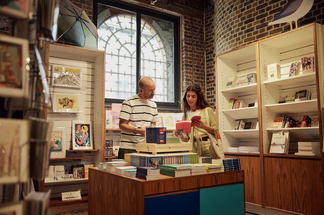 Two people browse books inside a bookstore, standing near wooden shelves filled with various books and other items, with a large window in the background.