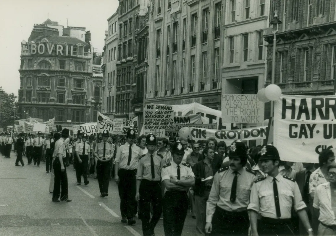 A black-and-white photo shows a march with participants holding signs advocating for LGBTQ+ rights. Police officers are seen in the front and along the sides of the march.