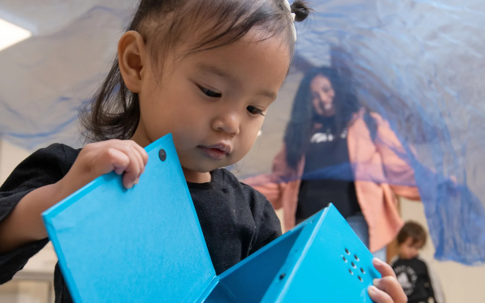 A toddler with pigtails examines a blue triangular toy in a playroom, with adults and another child visible in the background.