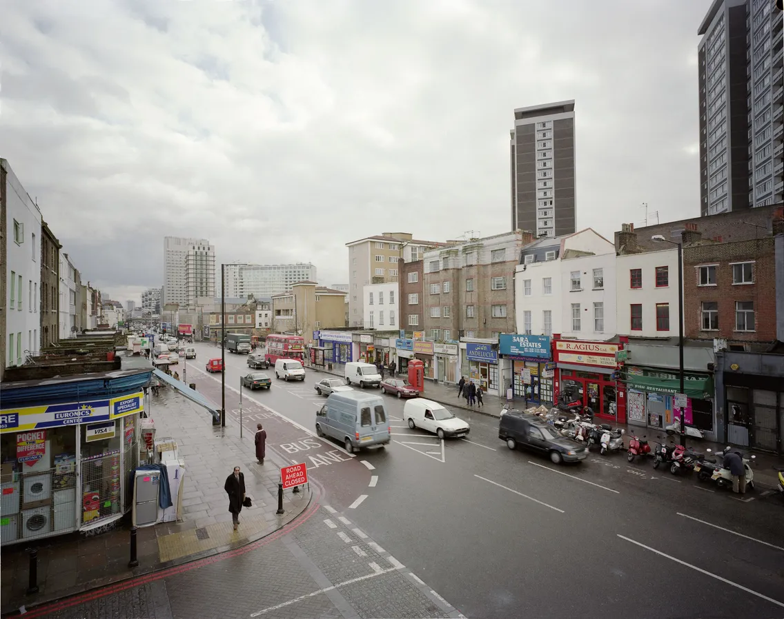A busy urban street with cars, pedestrians, and various shops under an overcast sky.