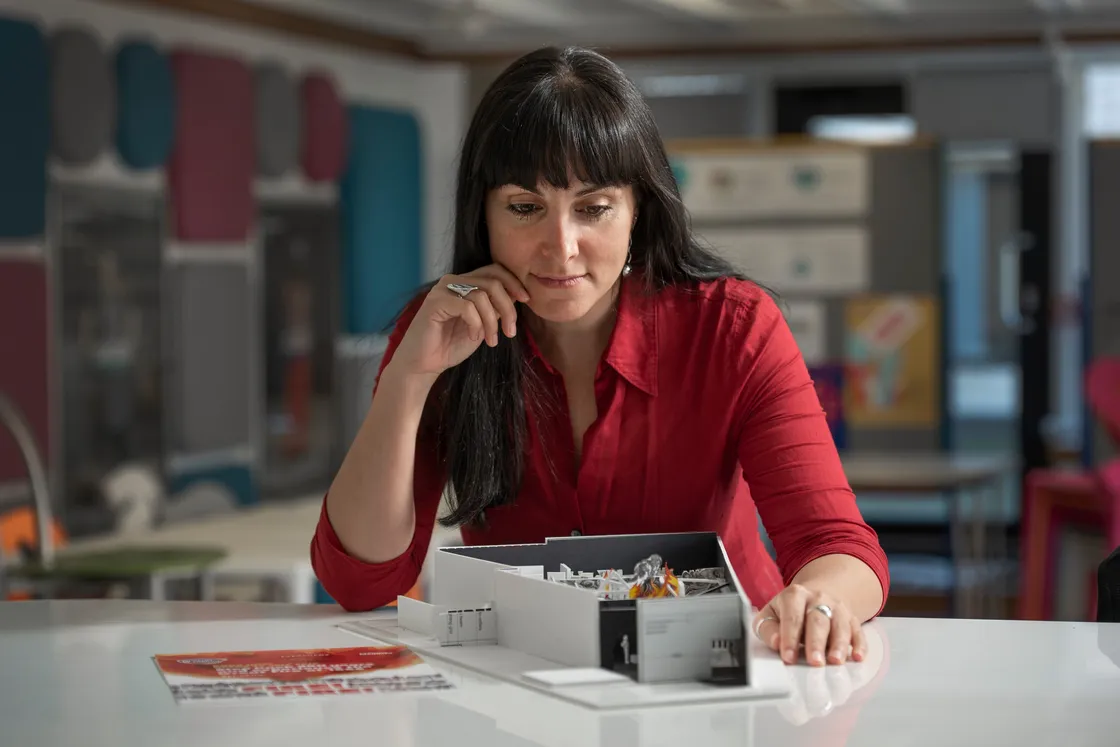 A woman in a red shirt is looking at a detailed architectural model on a table indoors. The background includes blurred architectural elements and other objects.