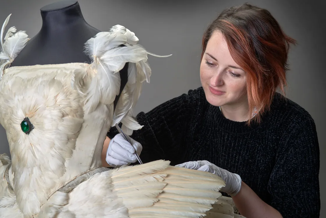A white woman wearing gloves tending to a white dress made predominantly of feathers