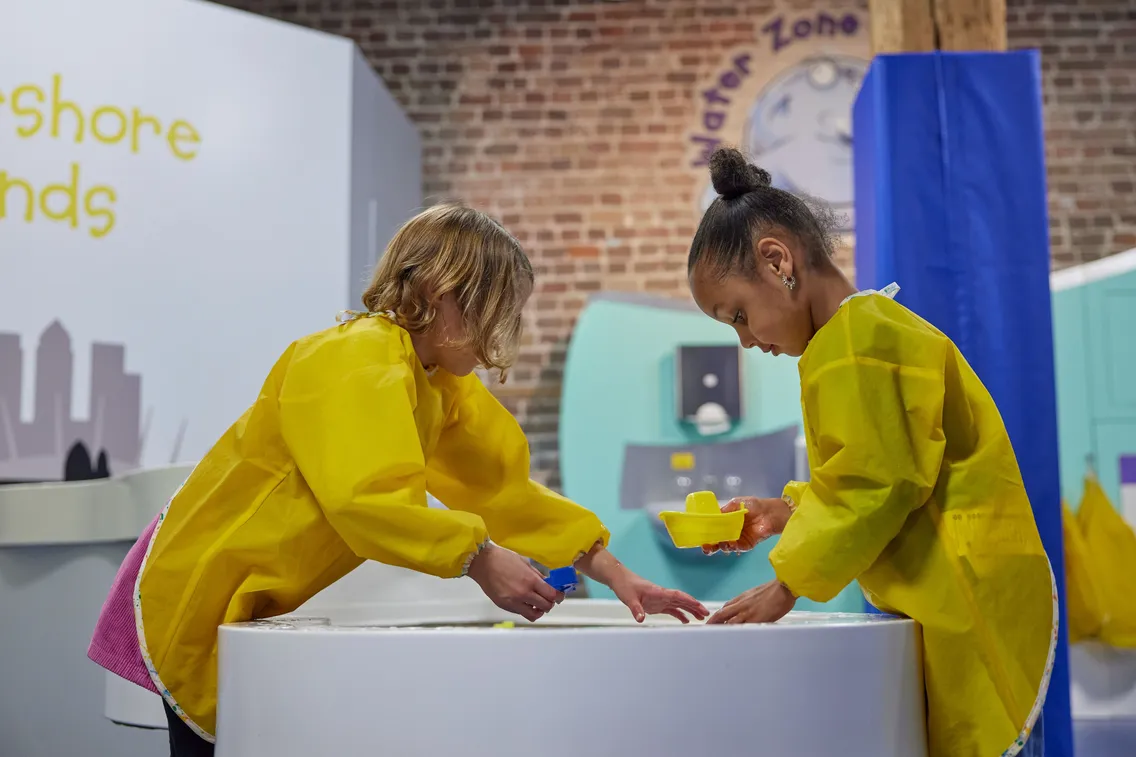 Two young girls wearing yellow overalls, playing at a water table