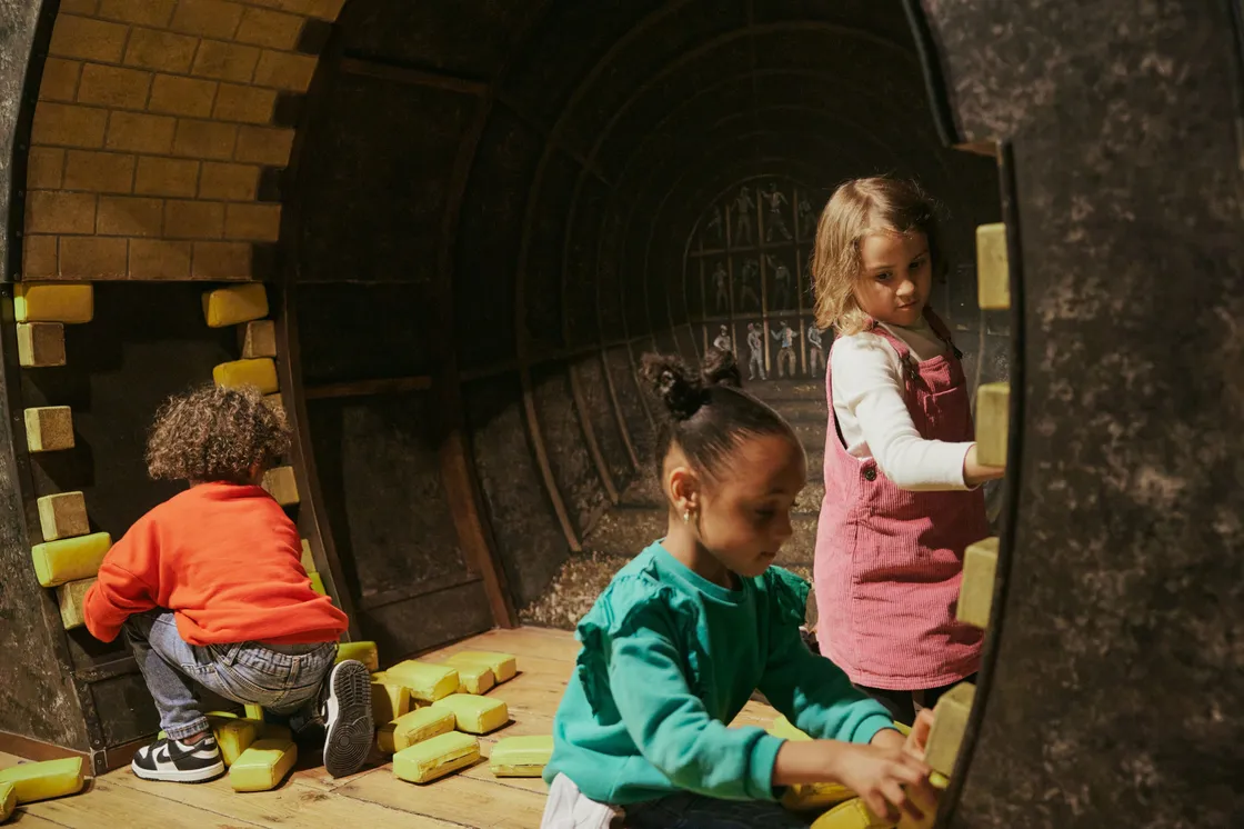 Three children in a recreation of a tunnel, piling up small yellow bricks