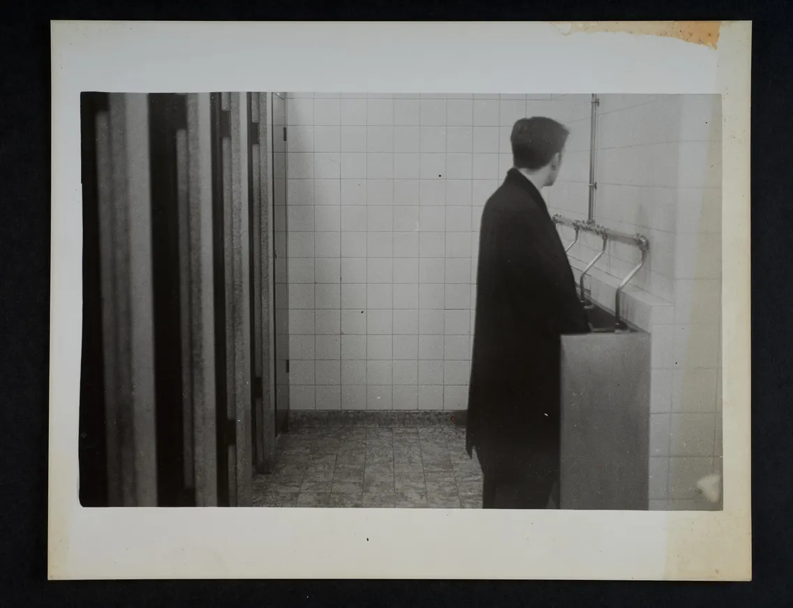 A man wearing a dark coat stands at a urinal in a tiled restroom, looking slightly to his right. The restroom has partitioned stalls on the left and a tiled floor.