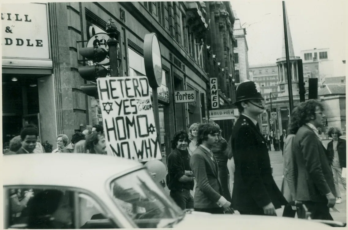 A group of people walk along a city street, one holding a sign reading "HETERO YES HOMO NO. WHY" A police officer is present, and cars are visible in the foreground.