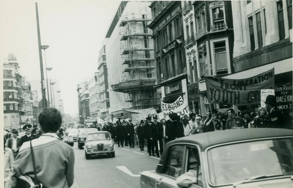 A black-and-white photo of a street protest with people holding signs and banners, including "Lesbians out" and "Youth group," while police officers line the street.