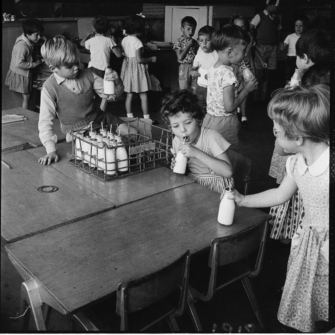 Children in a classroom drinking milk from glass bottles, with a metal crate of milk bottles on the table.