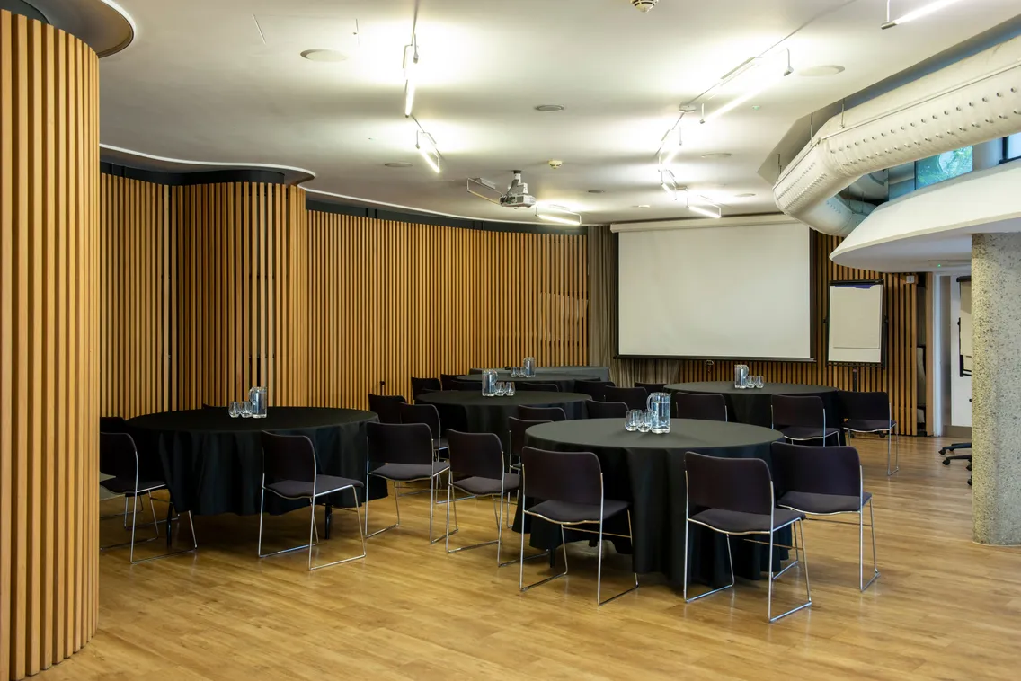 A conference room with round tables and chairs arranged, featuring a projector screen, flip chart, and water bottles on the tables. The room has wooden paneling and bright overhead lighting.