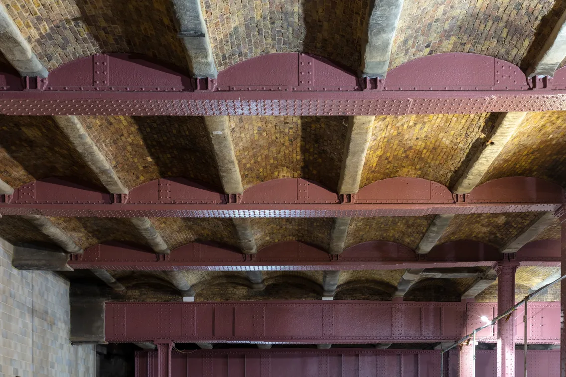 The image shows the interior of a building with a ceiling supported by arched steel beams and a roof constructed of bricks. The beams are painted red, and the walls are tiled.