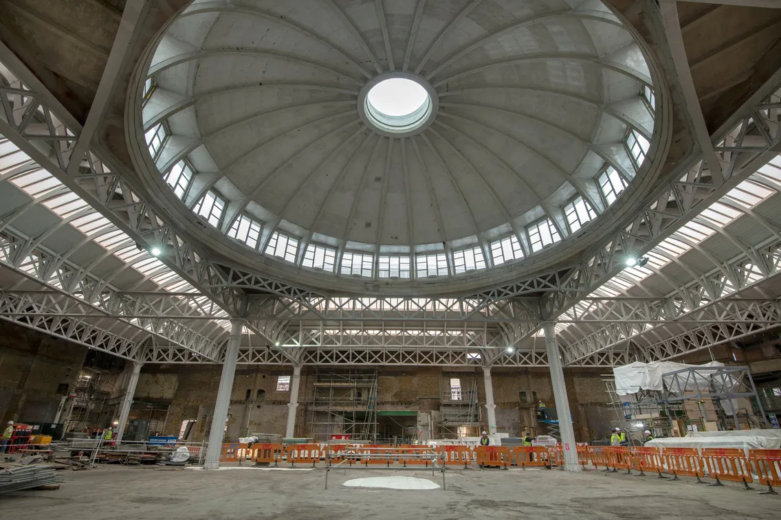 A large, empty industrial building under renovation with a circular dome ceiling, metal supports, and construction materials scattered around. Orange barriers cordon off parts of the site.