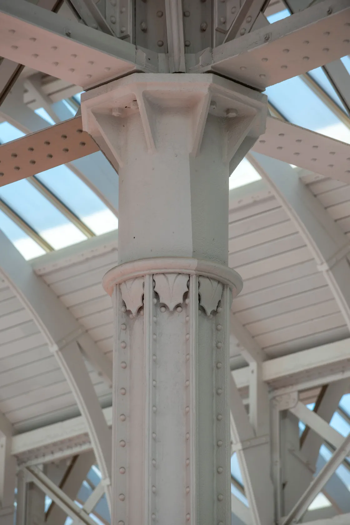 Close-up view of a sturdy structural column with rivets and decorative elements, supporting a roof with glass panels allowing natural light.