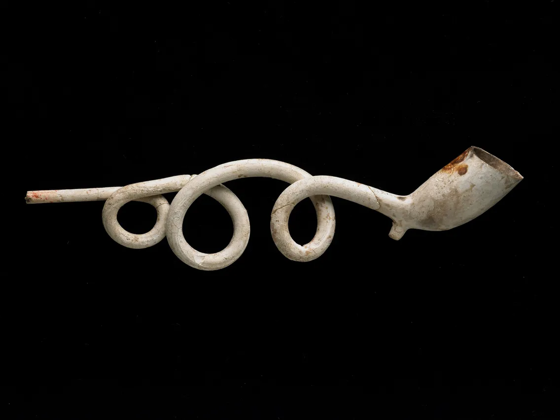 An old, white twisted clay pipe with a long stem and a bowl, set against a black background.