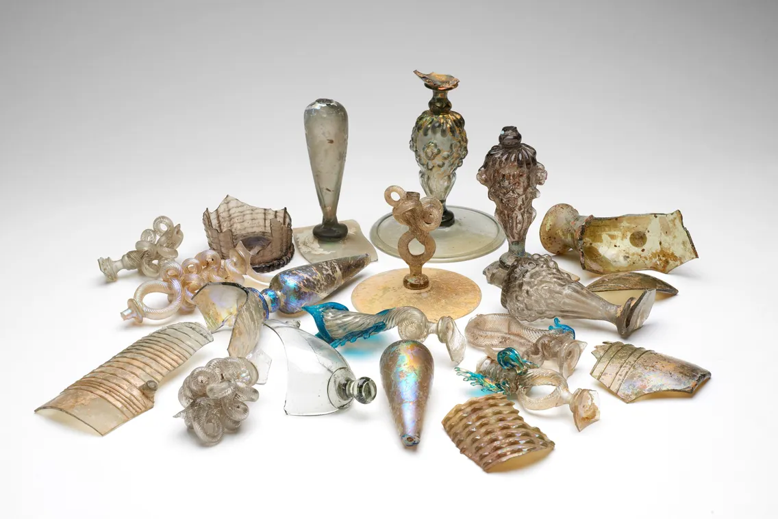 A collection of ancient glass artifacts of varying shapes and sizes, including vases, bottles, and decorative pieces, displayed against a plain background.