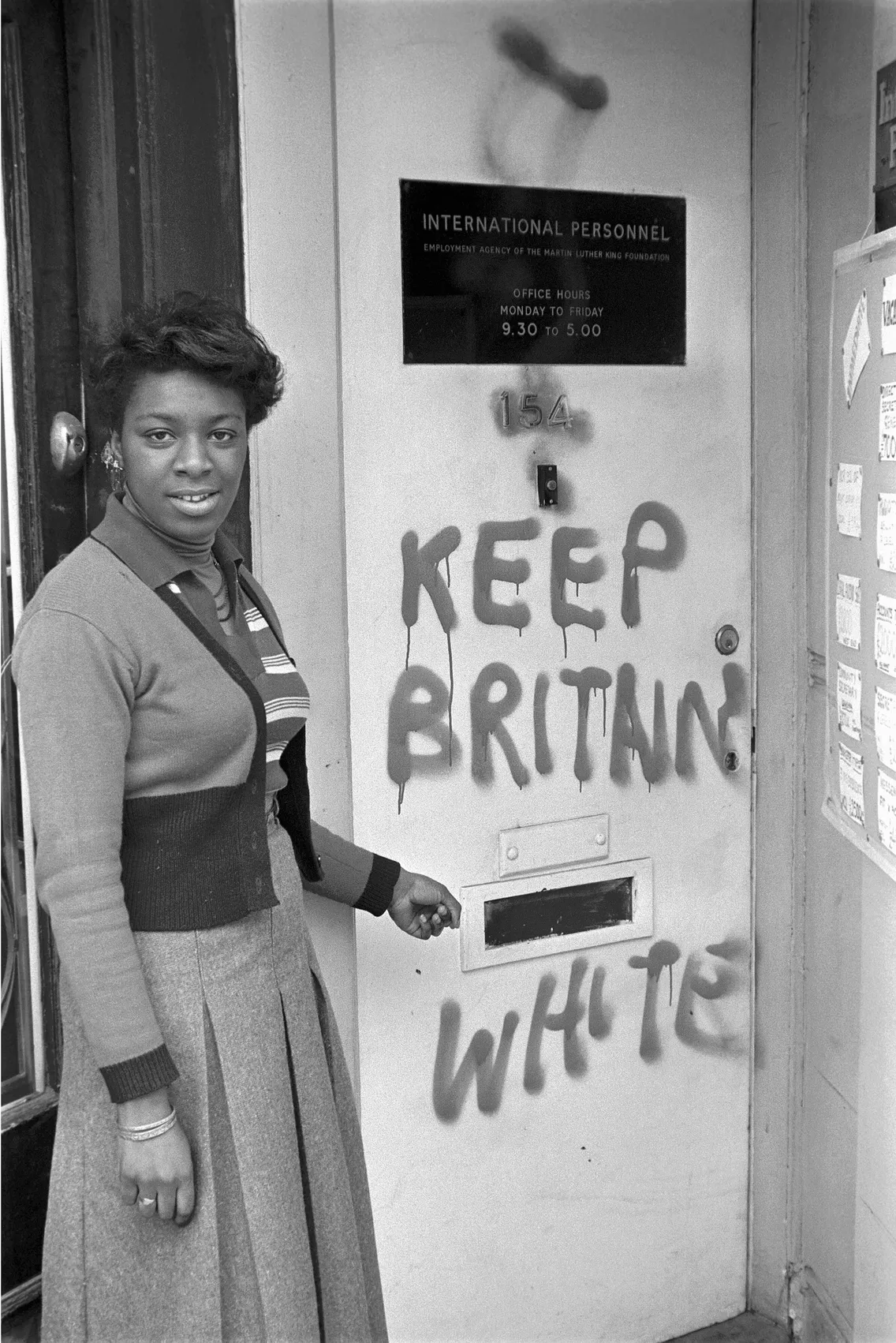 A woman stands by a door with "keep britain white" graffiti.