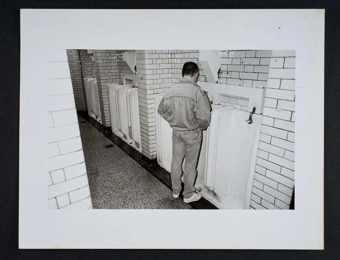 A man in a denim jacket and jeans using a urinal in a tiled public restroom. The image is in black and white.