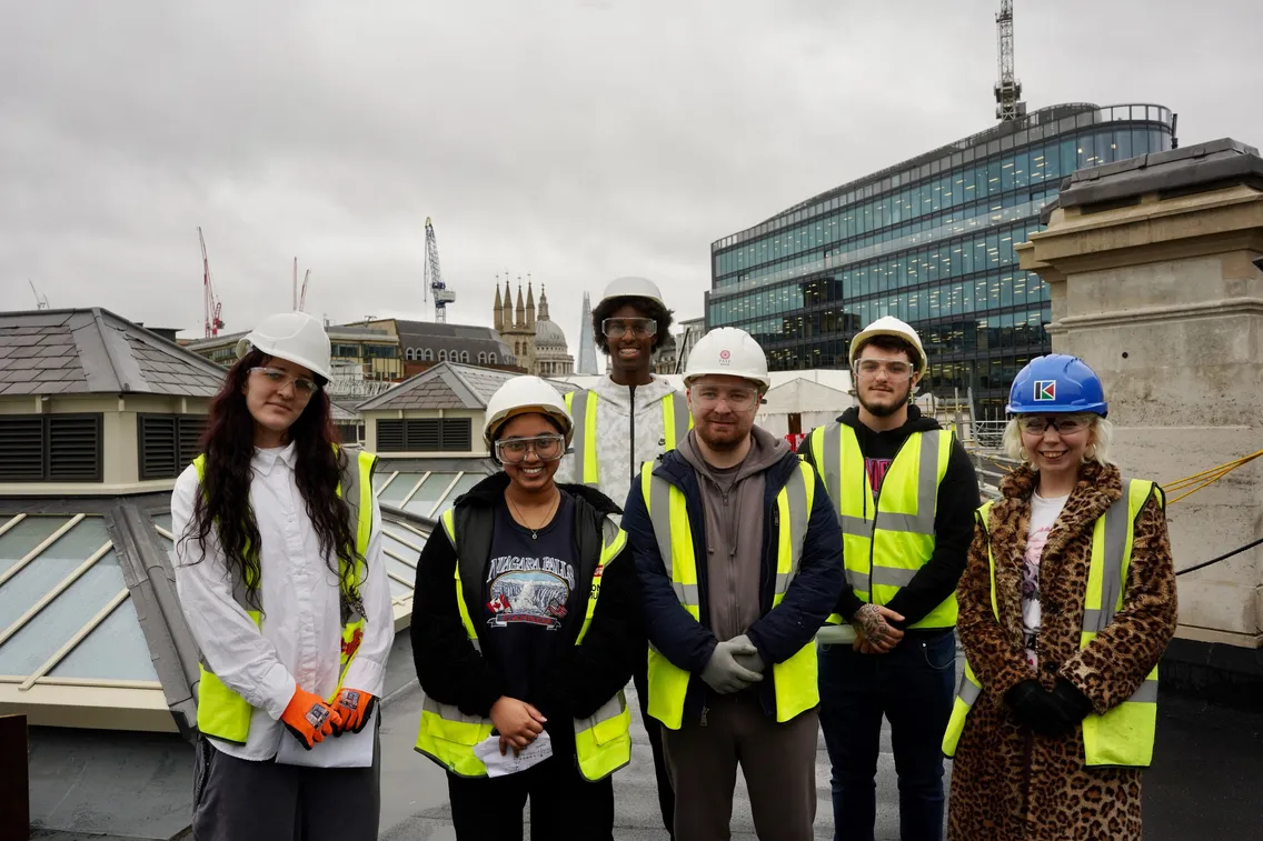 A group of six construction workers wearing safety gear, including helmets and high-visibility vests, standing on a rooftop with city buildings and cranes in the background.