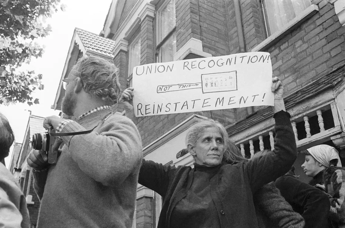 A person holds a sign demanding "union recognition" and "reinstatement" during a protest, with bystanders in the background.