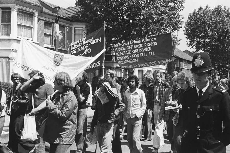 Black and white photo of a group of people demonstrating with banners about labor rights, escorted by a policeman, on a suburban street.