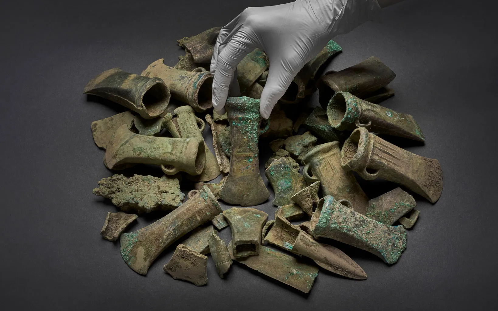 Two women are seated at a table with archaeological artifacts including a sword, a bronze spearhead, a hammer, and a flint tool. One woman holds up the sword, and a poster is in the background.