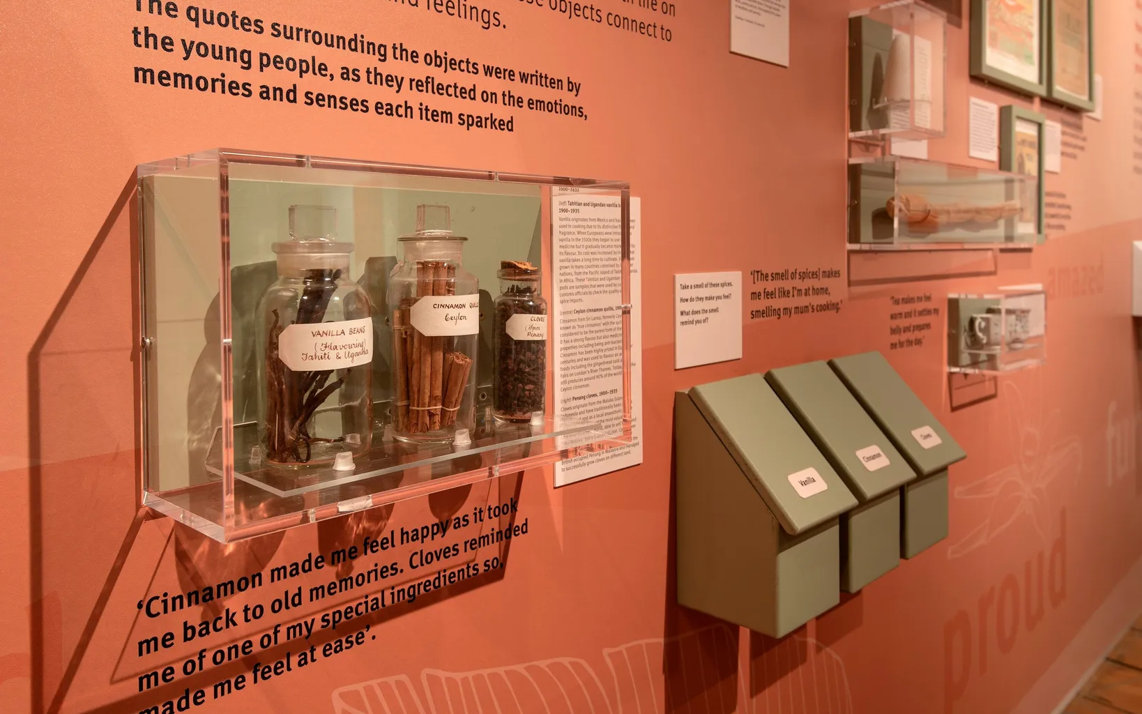 A display with labeled jars containing spices and herbs, alongside quotes about memories and feelings. There are also four green boxes affixed to the wall, presumably containing more items or information.