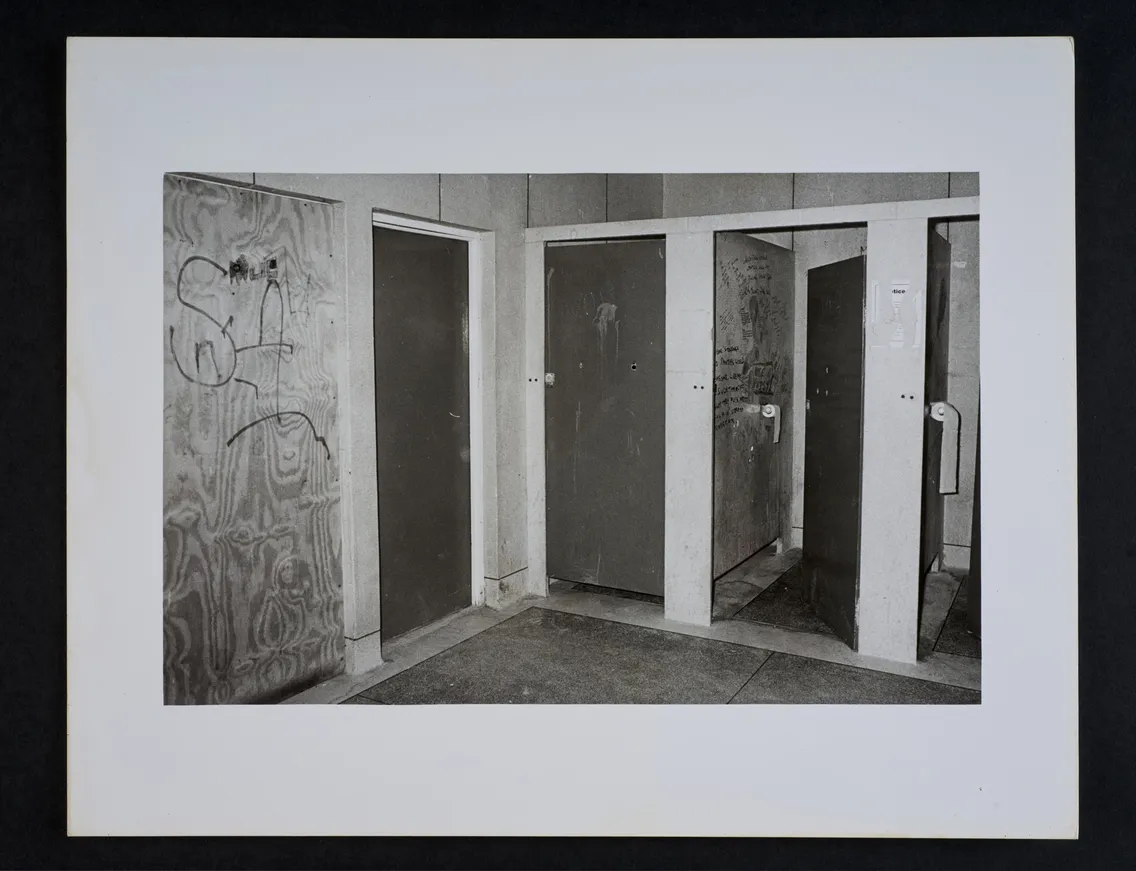 A black and white photograph of three bathroom stalls. The middle stall has a wooden door covered in graffiti, while the other two have solid metal doors. The area appears worn and unattended.