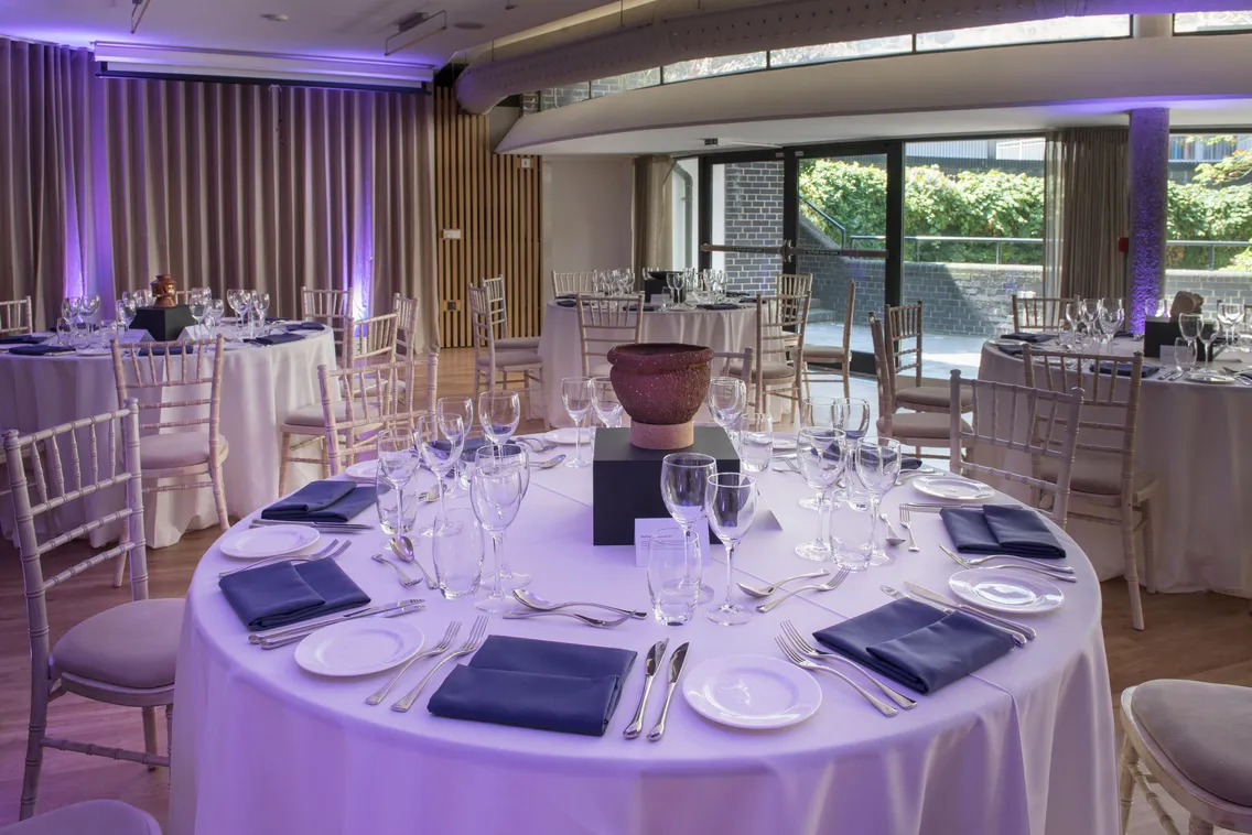 A banquet hall is set for an event with round tables covered in white tablecloths, navy blue napkins, and elegant glassware. Large windows and striped wooden paneling are in the background.