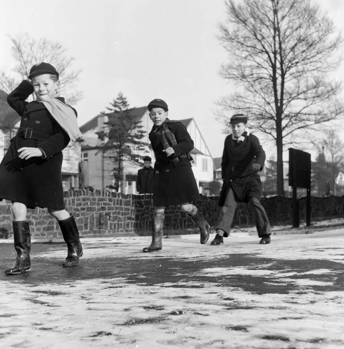 Three boys in winter attire, including coats and boots, joyfully skate on a frozen lake in a suburban area.