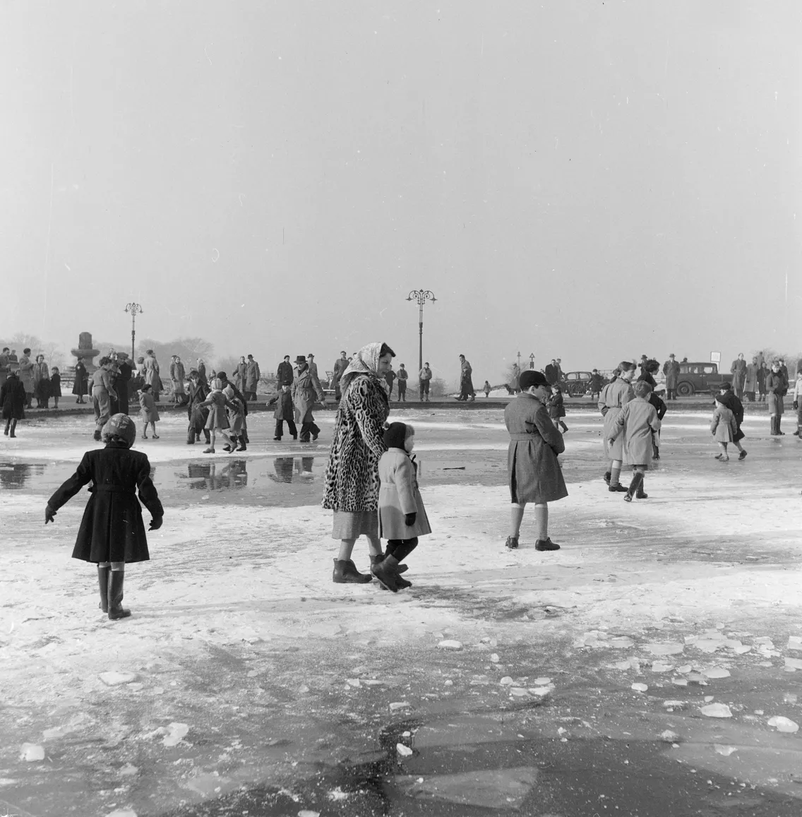 Black and white photo of people ice skating on a frozen outdoor rink, featuring a woman holding a child's hand, with other skaters and lamp posts in the background.