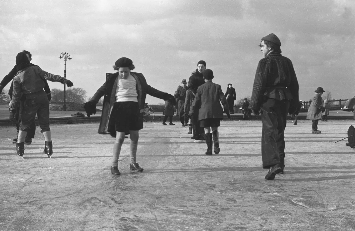 People ice skating on a frozen outdoor rink, with a girl in the foreground balancing as she skates and others scattered around the area.