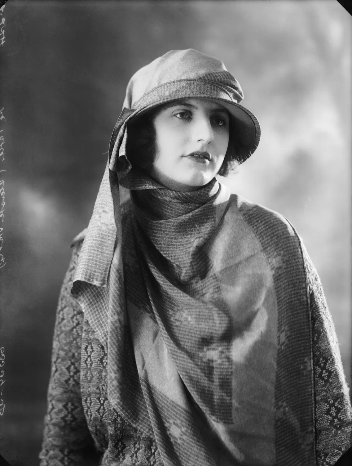 Black and white portrait of a woman wearing a cloche hat and draped in a patterned shawl, gazing to her right.