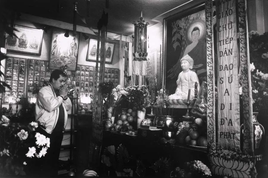 Man praying in a richly decorated buddhist shrine filled with fruit offerings, religious banners, and statues.