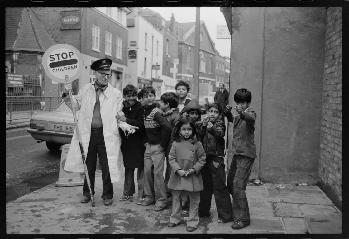 A black-and-white photo of a crossing guard holding a "stop children" sign, surrounded by a group of young children on a city street.
