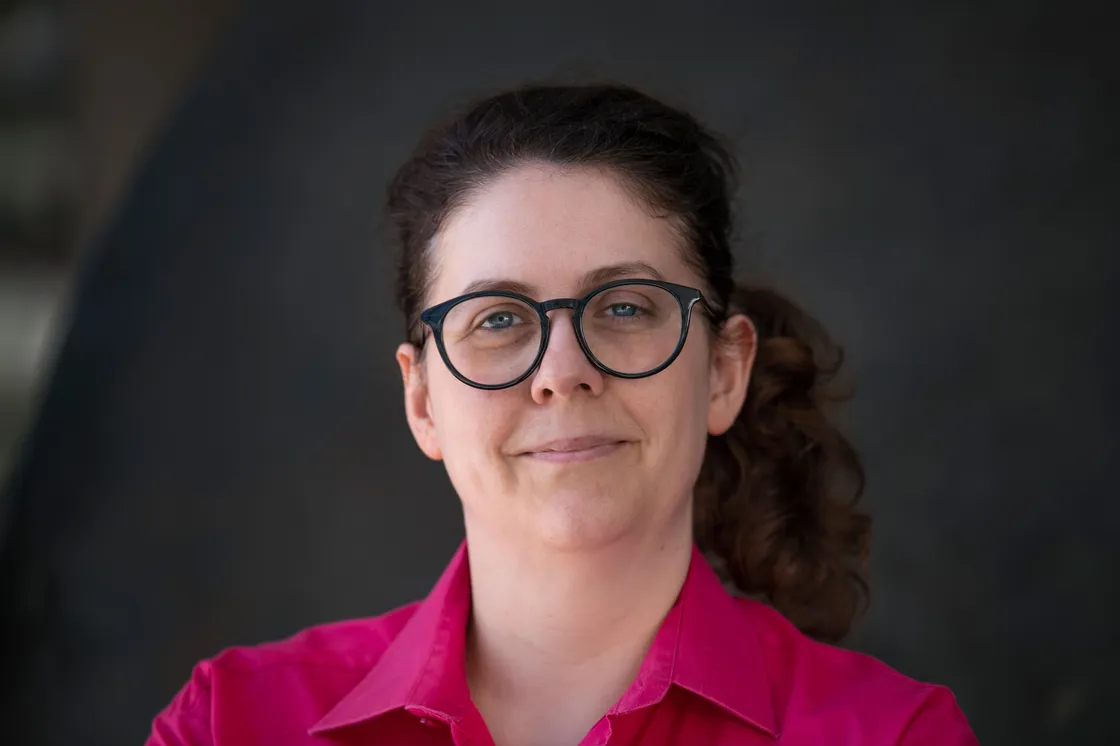 A white woman wearing with brown hair wearing glasses and a pink shirt, looking at the camera
