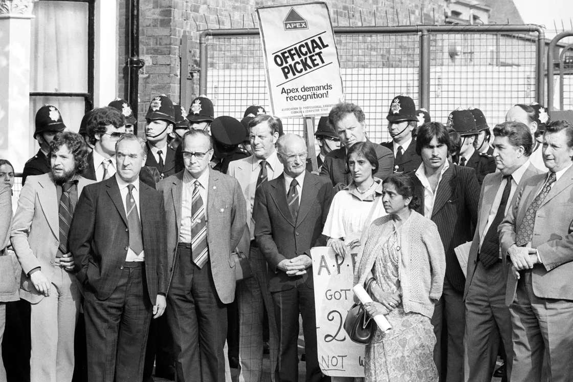 A group of people stands on the picket line in front of a gate with a sign reading "Official Picket" held by one person. Several police officers are visible behind them.